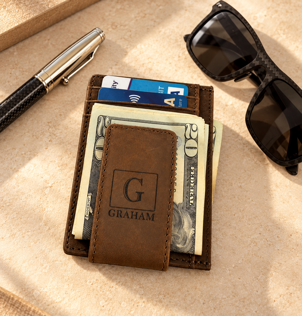 A slim brown leather wallet with a monogram 'Graham' inscribed on the front, displayed alongside sunglasses and a pen on a wooden surface.