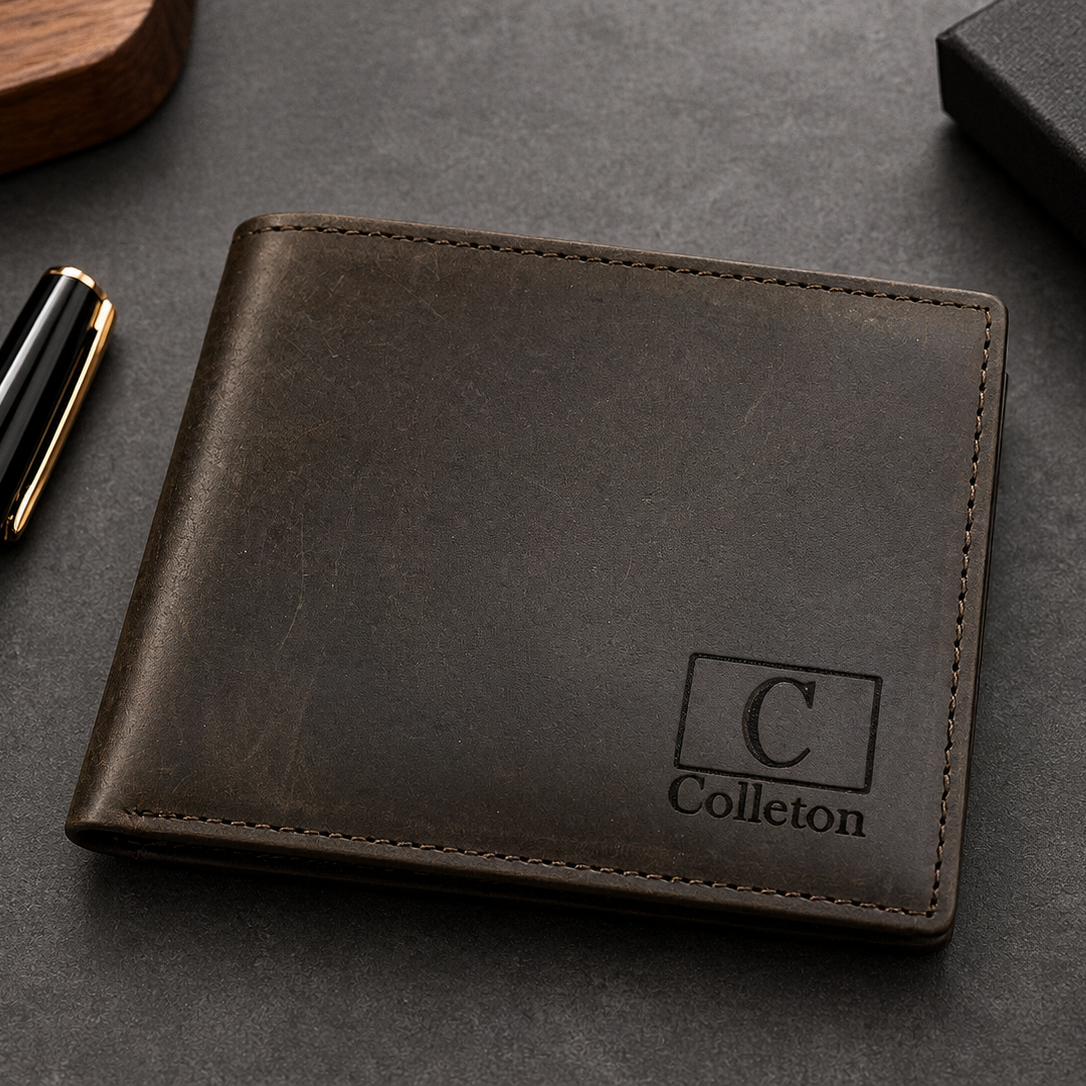 Personalized dark brown leather bifold wallet for men with engraved C monogram and Colleton name on the lower-right corner, displayed on a luxury matte charcoal desk with a black and gold pen. The lighthouse goods