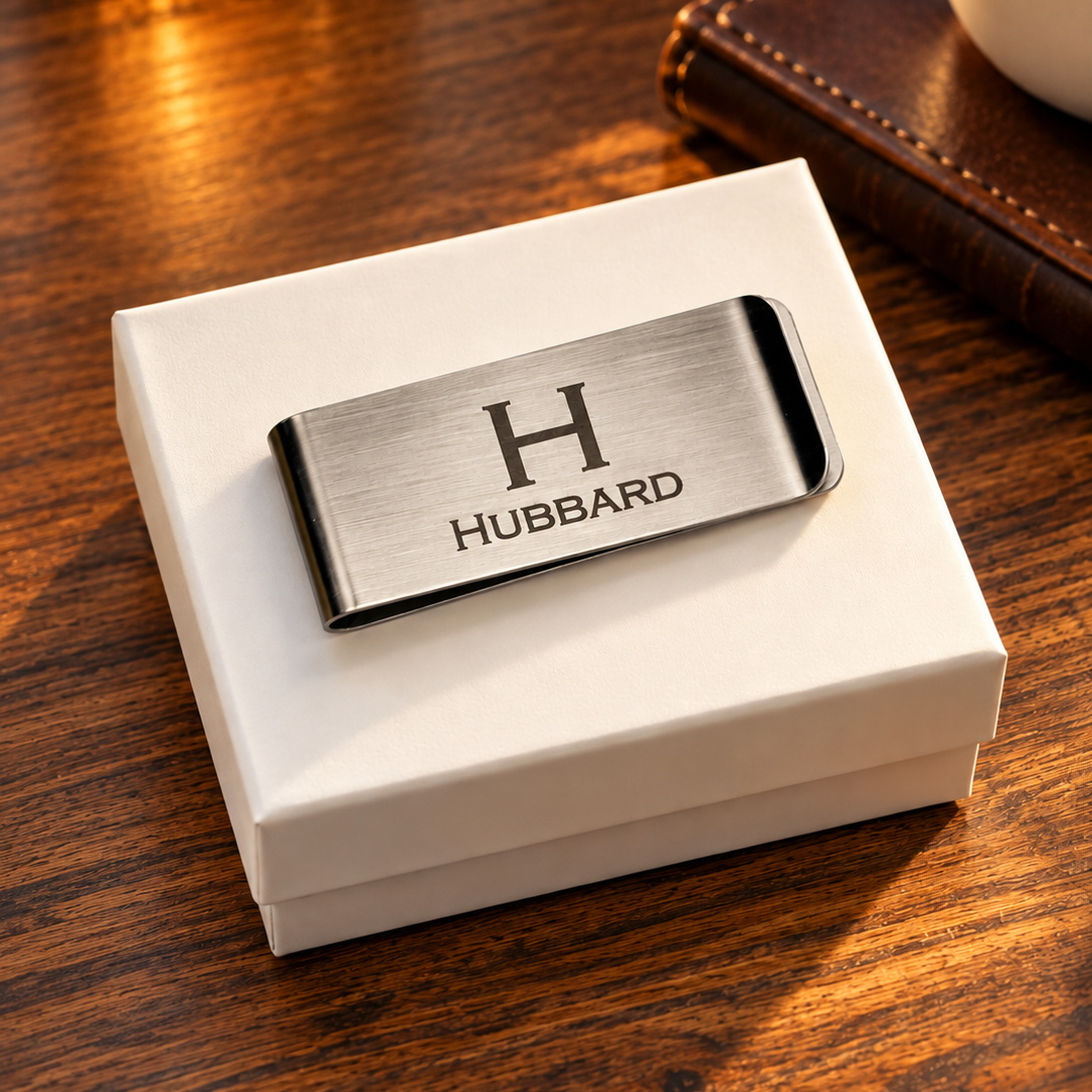 Personalized brushed stainless steel money clip engraved with H initial and HUBBARD name, displayed on a white gift box on a warm wooden desk.